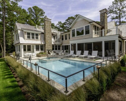 Modern two-story house with a stone chimney, large windows, and a spacious patio featuring an outdoor kitchen and a luxury pool, bordered by lounge chairs and landscaped greenery.