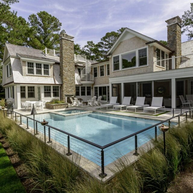 Modern two-story house with a stone chimney, large windows, and a spacious patio featuring an outdoor kitchen and a luxury pool, bordered by lounge chairs and landscaped greenery.