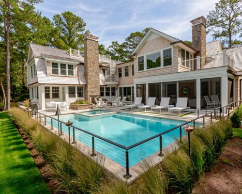 Large modern house with stone chimneys and multiple windows, featuring an inground pool, lounge chairs, and landscaped garden in a wooded area—perfect for outdoor living.