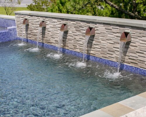 Rectangular inground pool with blue tile and a stone wall featuring five metal spouts, each with water flowing into the pool below.