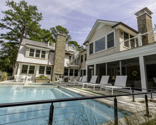 Large modern house with stone chimneys, multiple windows, and a fenced outdoor patio designed for outdoor living, featuring an inground fiberglass pool and stylish white lounge chairs.