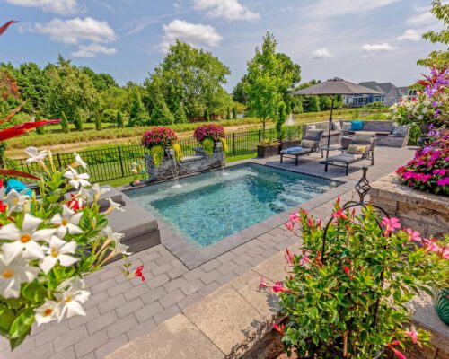 A backyard with a small rectangular fiberglass pool, patio seating, umbrella, potted flowers, and greenery in the background under a partly cloudy sky.