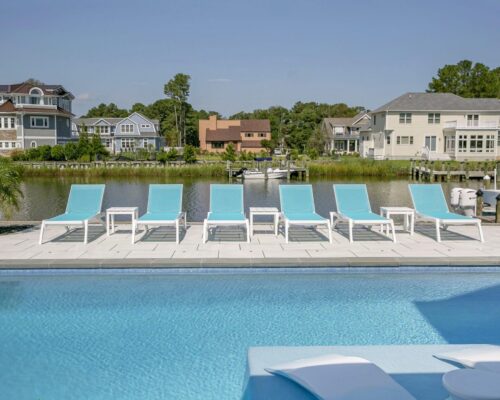 Row of blue lounge chairs and white tables beside a luxury pool, overlooking a canal with houses and trees in the background under a clear sky—perfect for outdoor living.