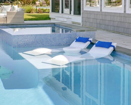 Two white lounge chairs with blue pillows and a small round table are partially submerged in a luxury pool next to a raised hot tub, creating the ultimate outdoor living and relaxation space.