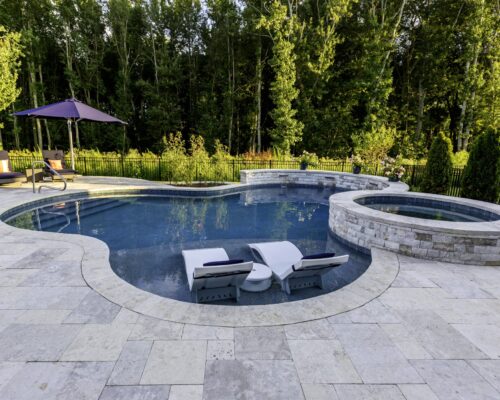 Backyard fiberglass pool with two loungers partially submerged in water, adjacent to a circular hot tub, surrounded by a stone patio and trees—perfect for outdoor living.