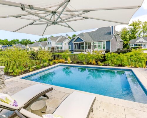 Rectangular backyard fiberglass pool with two lounge chairs, umbrella, and landscaping, surrounded by a fence, with houses in the background—perfect for outdoor living.