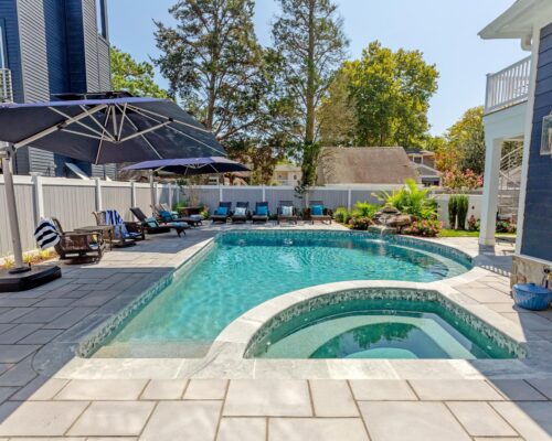 A backyard luxury pool with clear water, surrounded by a stone patio, lounge chairs, large umbrellas, and landscaping next to a blue house and white fence—perfect for those seeking premium inground pools.