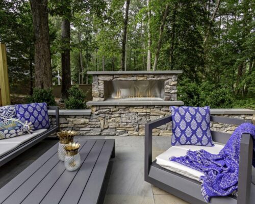 Outdoor patio with gray cushioned seating, patterned blue pillows, a blue throw blanket, a stone fireplace, and views of lush trees—perfect for relaxing after a swim in nearby luxury pools or custom spas.