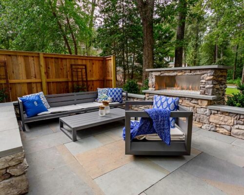 Modern outdoor patio with gray furniture, blue patterned cushions, a stone fireplace, and a wooden fence surrounded by trees—perfect for relaxing after enjoying custom spas or elegant outdoor kitchens nearby.
