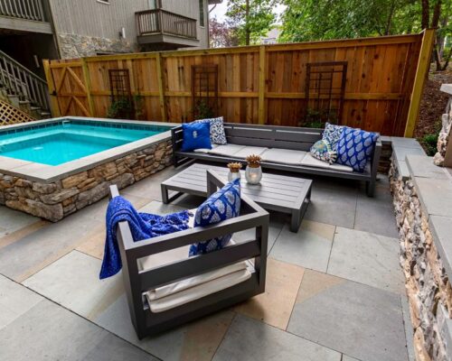 A small backyard patio with gray seating, a rectangular coffee table, stone accents, and a luxury pool surrounded by a wooden fence and greenery.