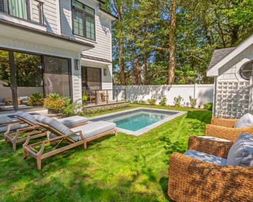 A modern backyard featuring a small rectangular fiberglass pool, lounge chairs, wicker armchairs, and a shed, all surrounded by grass, plants, and a white fence.