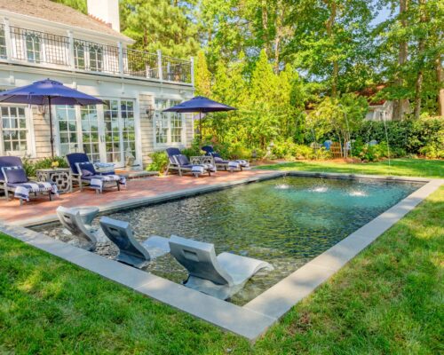 Rectangular backyard luxury pool with three built-in loungers, surrounded by patio chairs, umbrellas, and greenery beside a house with large windows—perfect for relaxing in style.