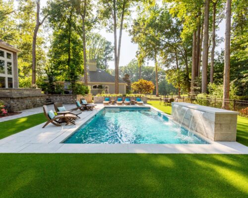 Rectangular backyard fiberglass pool with lounge chairs, a stone waterfall feature, and surrounding artificial grass, set among tall trees and adjacent to a house. Perfectly complements spaces with outdoor kitchens or custom spas.