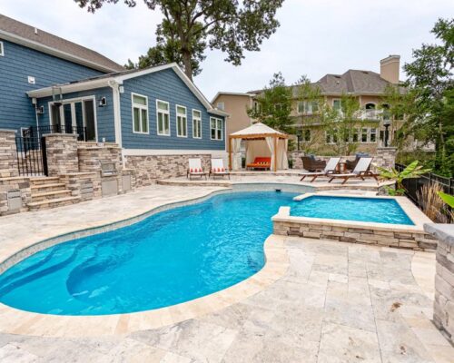 Backyard with a stone patio, fiberglass pool, hot tub, lounge chairs, and a cabana, adjacent to a blue house and surrounded by greenery—perfect for relaxing or entertaining with ample space for outdoor kitchens.