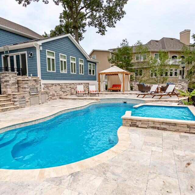 Backyard with a stone patio, fiberglass pool, hot tub, lounge chairs, and a cabana, adjacent to a blue house and surrounded by greenery—perfect for relaxing or entertaining with ample space for outdoor kitchens.