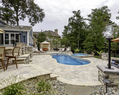 Stone patio with inground pools and hot tub, surrounded by lounge chairs, outdoor kitchen, bar area, and trees; house in the background under a cloudy sky.