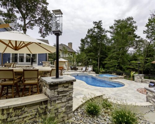 Backyard patio with stone flooring, large dining table under an umbrella, inground pool and custom spa surrounded by trees and outdoor living seating areas.