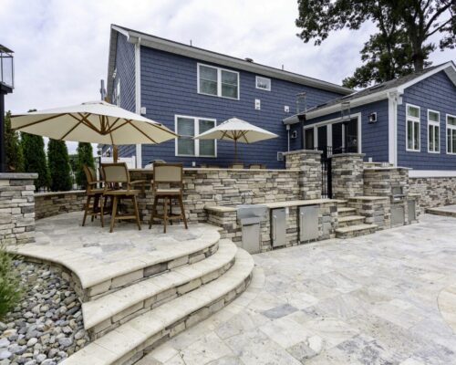 A modern backyard patio with stone steps, outdoor seating, large umbrellas, and a blue house exterior, adjacent to luxury pools—perfect for elevated outdoor living.