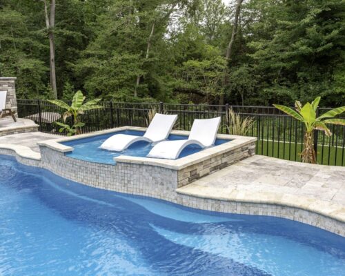 Two white lounge chairs sit partially submerged in a small elevated fiberglass pool adjoining a larger swimming pool, surrounded by stone tile and trees in the background.