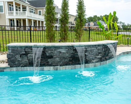 A swimming pool with three stone waterfalls, clear blue water, and a black metal fence sits near a residential area—perfect for enjoying inground pools or planning custom spas and outdoor kitchens nearby.