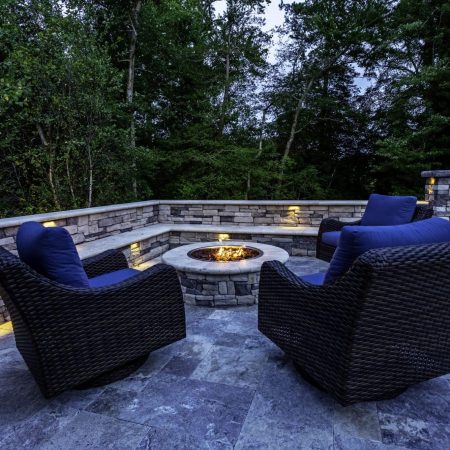 Stone patio with three wicker chairs and a fire pit in the center, surrounded by stone walls and outdoor lighting, set against a backdrop of trees—perfect for relaxing after a swim in nearby luxury pools or enjoying evenings by custom spas.