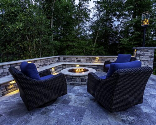 Stone patio with three wicker chairs and a fire pit in the center, surrounded by stone walls and outdoor lighting, set against a backdrop of trees—perfect for relaxing after a swim in nearby luxury pools or enjoying evenings by custom spas.