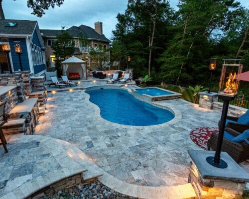 Stone patio backyard with a fiberglass pool, hot tub, lounge chairs, fire pit, gazebo, bar seating, and surrounding trees at dusk—perfect for outdoor living.