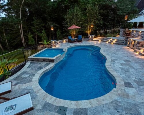 A backyard features a kidney-shaped luxury pool with an attached hot tub, lounge chairs on a stone patio, surrounding trees, outdoor lighting, and space for outdoor kitchens.