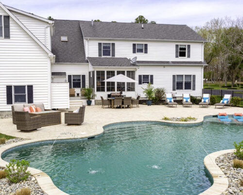 A large backyard features an inground swimming pool with lounge chairs, a patio seating area, custom spas, and a white two-story house in the background.