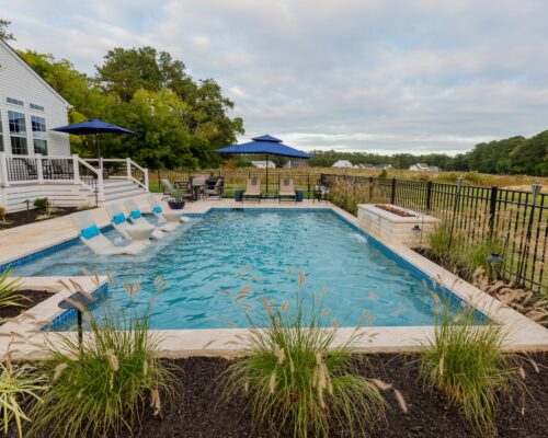 Rectangular inground pool with built-in loungers, a raised hot tub, patio seating, black metal fence, and landscaping—perfect for outdoor living—adjacent to a house and open grassy field.