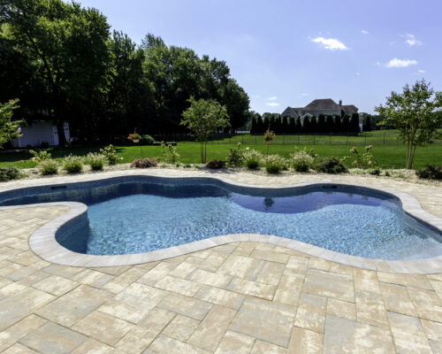 Backyard swimming pool with a curved shape, surrounded by a paved deck, landscaped garden beds, and lawn—this inviting space features luxury pools with outdoor kitchens nearby, all set amid trees and a house under a clear sky.