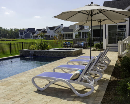 Four white lounge chairs with blue cushions line the side of a rectangular fiberglass pool, with a large umbrella and suburban houses in the background—perfect for relaxing or enjoying outdoor living.