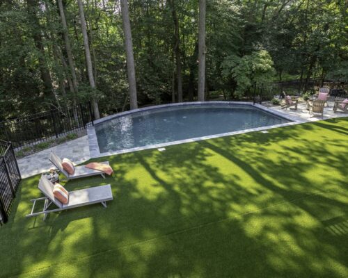 A backyard with a fiberglass pool, artificial turf lawn, lounge chairs, and a fire pit seating area, surrounded by trees and a black metal fence—perfect for outdoor living.