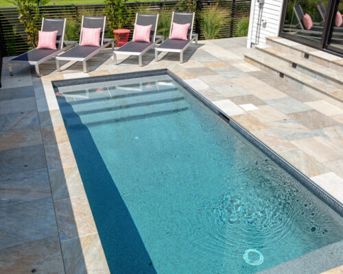 A small rectangular luxury pool with clear water is surrounded by stone tiles. Four lounge chairs with pink cushions are lined up along one side, creating an inviting backyard retreat.