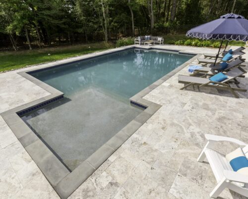 Rectangular inground pool with a shallow ledge, surrounded by lounge chairs, a patio umbrella, and green lawn bordered by trees—perfect for elevated outdoor living.