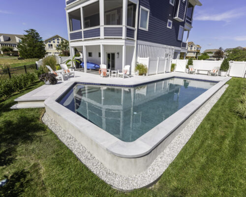 A modern rectangular inground pool is surrounded by a concrete deck, lounge chairs, and grass, creating the perfect outdoor living space in the backyard of a two-story house.