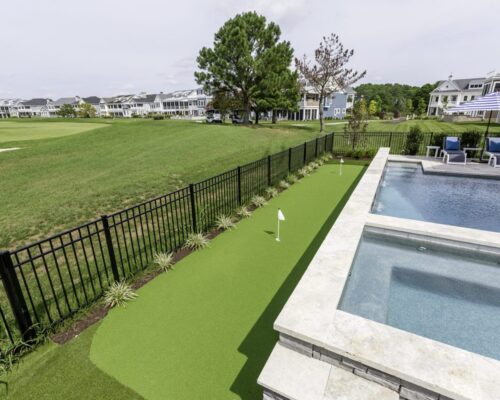 View of a backyard with a small artificial putting green beside a fenced luxury pool area, overlooking a golf course and residential homes in the background.