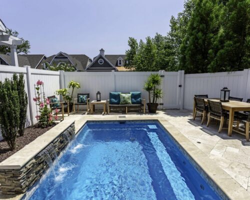 A small backyard with a rectangular fiberglass pool, stone deck, outdoor seating, dining table, plants, and a white privacy fence—perfect for outdoor living.