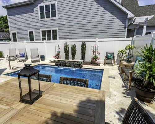 A small backyard with a rectangular luxury pool, lounge chairs, potted plants, a wooden dining table, and a tall white privacy fence creates the perfect outdoor living retreat.