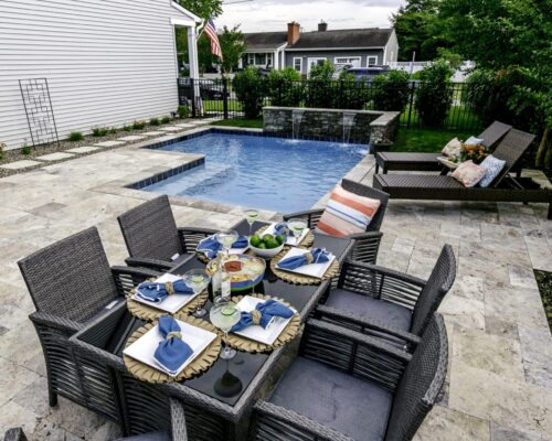 Outdoor patio with a glass-top dining table set for six, a small inground pool with a jacuzzi, and two lounge chairs, surrounded by a stone tile deck and fenced yard.