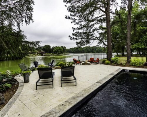 Two lounge chairs face a lake on a stone patio beside a luxury pool, surrounded by trees, greenery, and inviting outdoor living spaces.