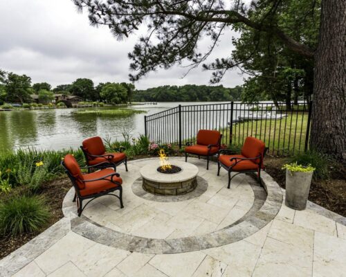 Four red cushioned chairs surround a fire pit on a stone patio beside a lake, with trees, a metal fence, and views perfect for enjoying nearby luxury pools or custom spas in the tranquil setting.