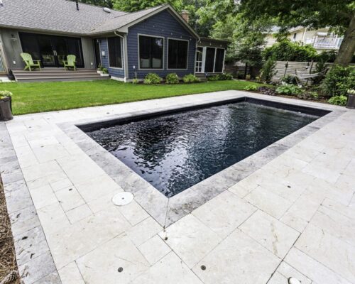 Rectangular inground pool surrounded by light stone tiles, with a lawn, potted plants, and a gray house featuring a patio and green chairs in the background—perfect for stylish outdoor living.