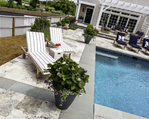 Two striped lounge chairs with a small table sit beside an inground pool on a stone patio, surrounded by potted plants and a house with a pergola in the background—perfect for outdoor living.