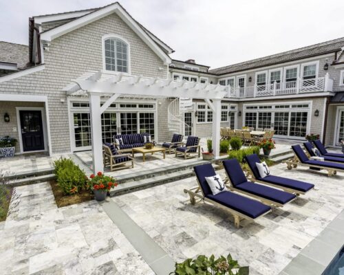 Backyard view of a large modern house with a pergola, outdoor seating area, dining table, and blue cushioned lounge chairs beside an inground pool on a stone patio—perfect for enjoying outdoor living.
