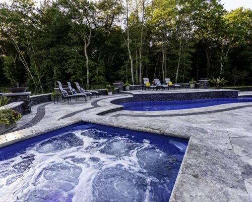A bubbling hot tub in the foreground overlooks a curving inground pool surrounded by lounge chairs and dense green trees, perfect for outdoor living.