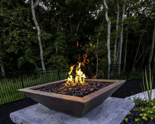 Square fire pit with flames sits on a stone pedestal in an outdoor garden area, complemented by views of luxury pools, with trees and a flower pot visible in the background.