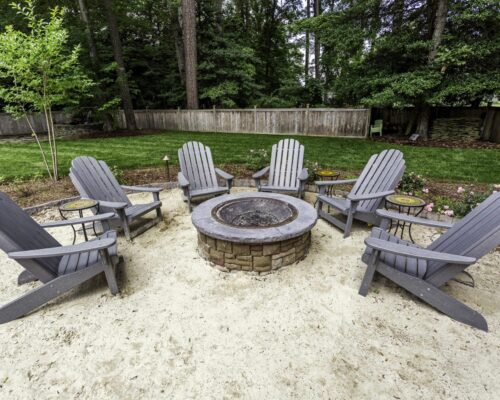 Six gray Adirondack chairs circle a stone fire pit in a fenced backyard with trees and grass, creating the perfect spot to relax after enjoying nearby outdoor kitchens or luxury pools.