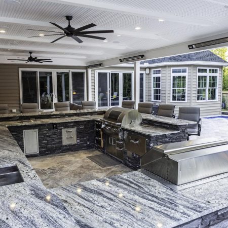 Modern outdoor kitchen with granite countertops, stainless steel appliances, ceiling fans, and a seating area—perfect for outdoor living—overlooks a pool, custom spas, and a gazebo in the background.