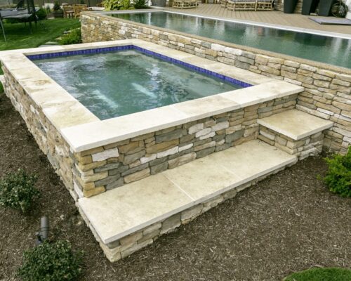 A small, square stone hot tub with clear water sits beside a raised swimming pool, surrounded by landscaped greenery and mulch—perfect for custom spas and enhancing your outdoor living experience.
